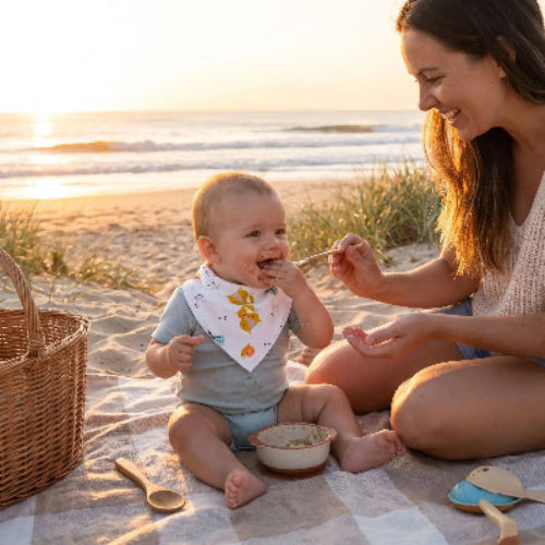 Bavoir bandana au cou de bébé assis sur une couverture et maman lui donne à manger prêt de la mer
